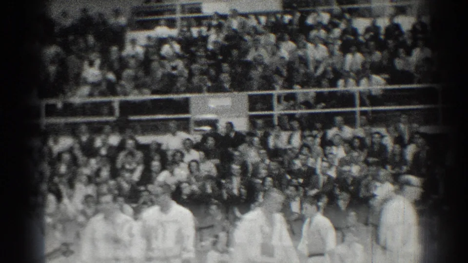 Black-and-white 8mm frame from San Gabriel showing gi-clad students in the foreground and a dense gym audience in the bleachers.
