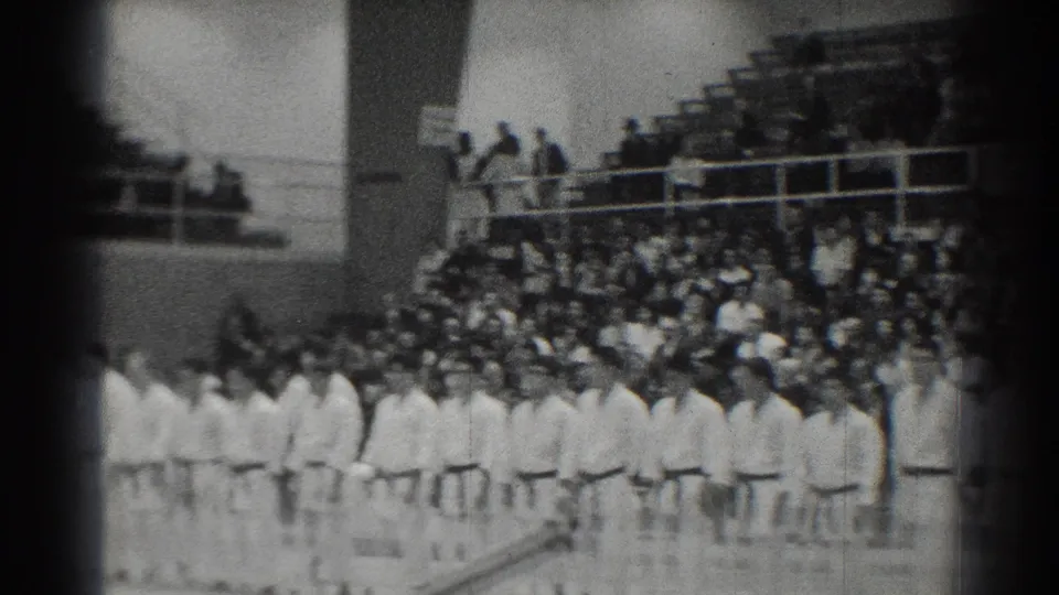 1966 San Gabriel home-movie frame showing a formal line of martial arts students in white gi facing across an indoor gym with spectators nearby.