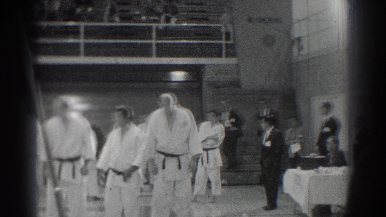 Several martial artists stand near suited officials and a covered table inside a gym.