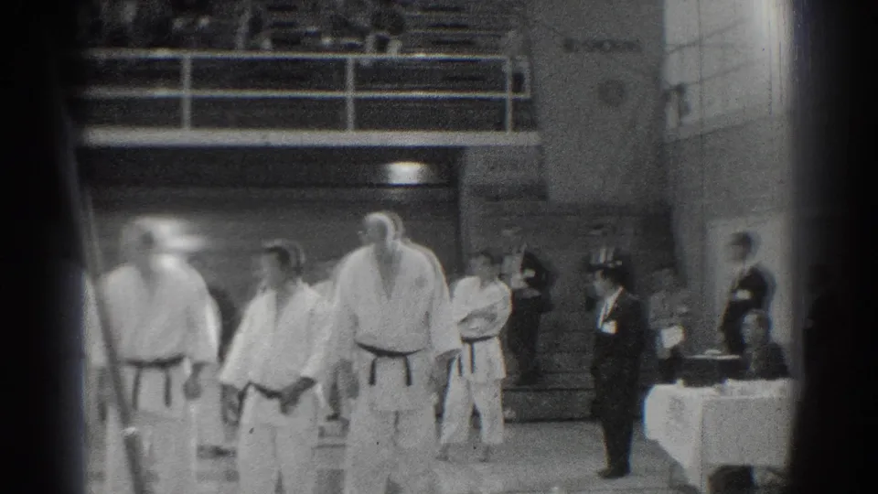 Martial artists in white uniforms stand beside officials in suits during the San Gabriel competition footage.