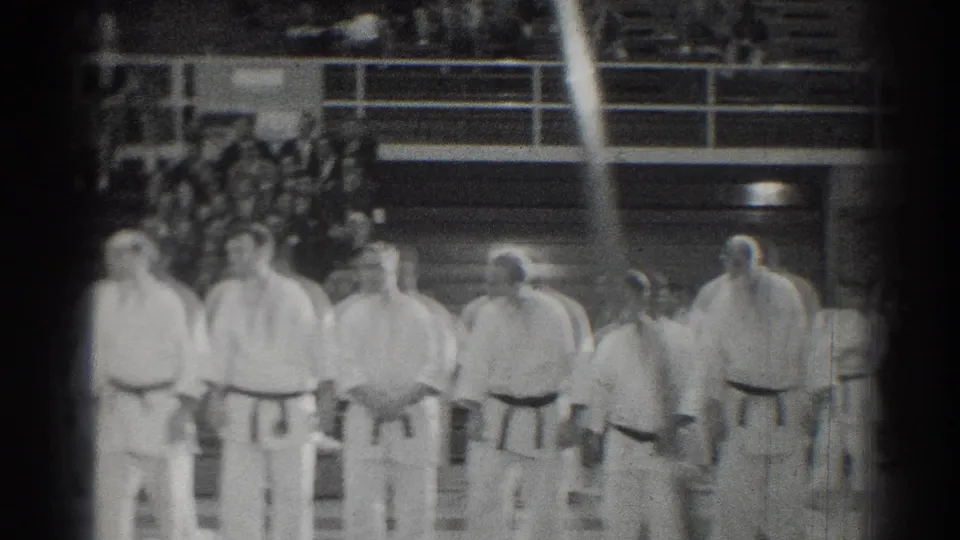 A row of uniformed martial artists faces the gym floor as a seated crowd watches from the stands.