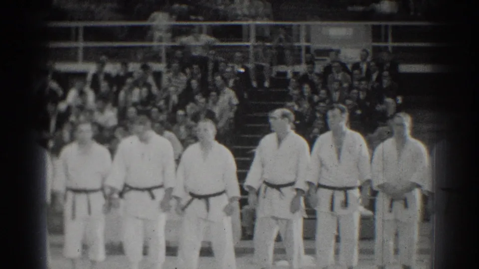 San Gabriel martial arts competitors stand in formation before a packed gymnasium audience in black-and-white 8mm footage.