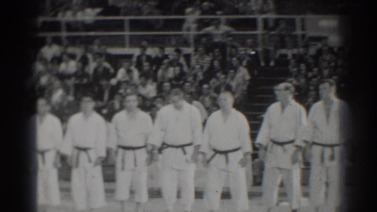 In San Gabriel, California in 1966, male karate competitors in white gi and dark belts stand at attention before a seated crowd.