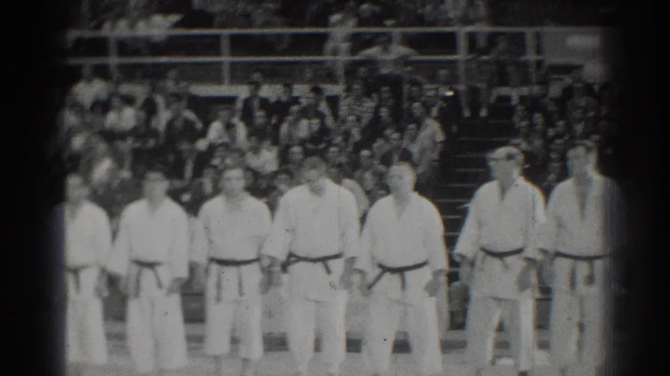 In San Gabriel, California in 1966, male karate competitors in white gi and dark belts stand at attention before a seated crowd.