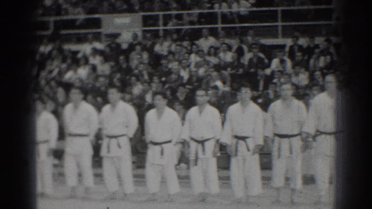 A row of karate competitors stands at attention with a large audience behind them.