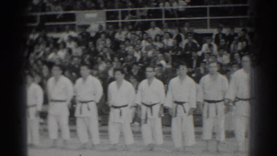 Male karate competitors stand in formation during the 1966 San Gabriel tournament, with spectators seated closely behind them.