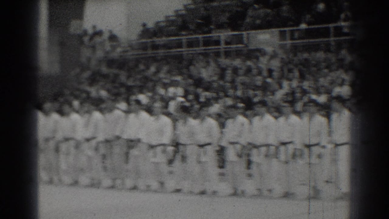 Martial artists in white gi stand in rows before a crowded bleacher section.