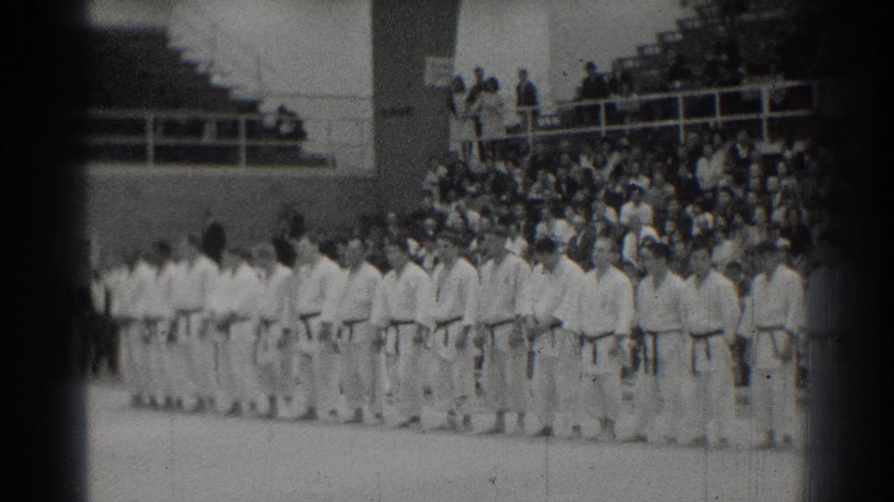 A long row of martial artists in gi stands across the floor near packed bleachers.
