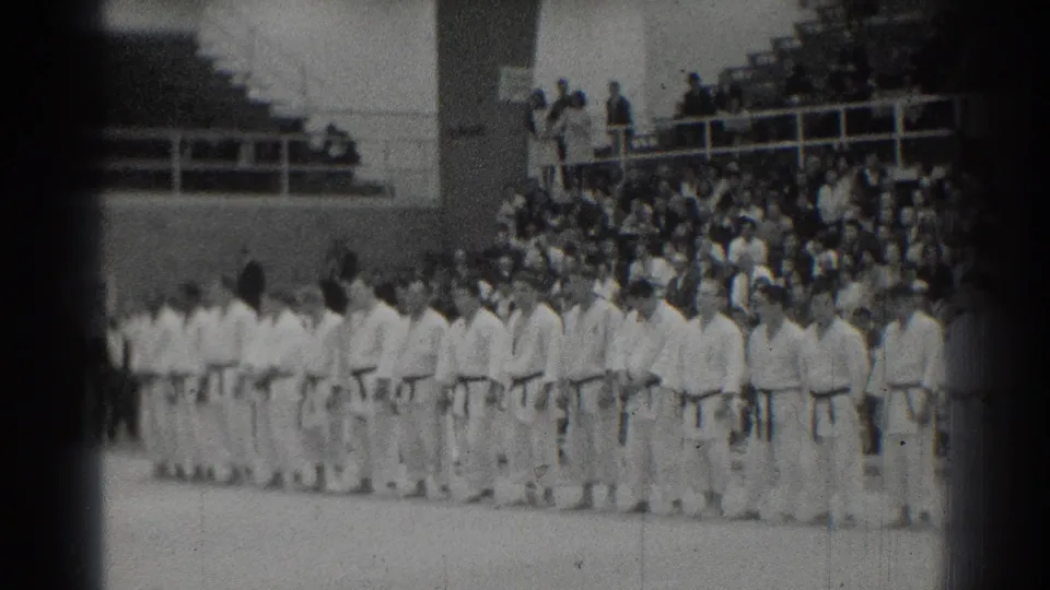 A line of martial arts competitors in white gi stands across a San Gabriel competition floor with spectators in the stands.