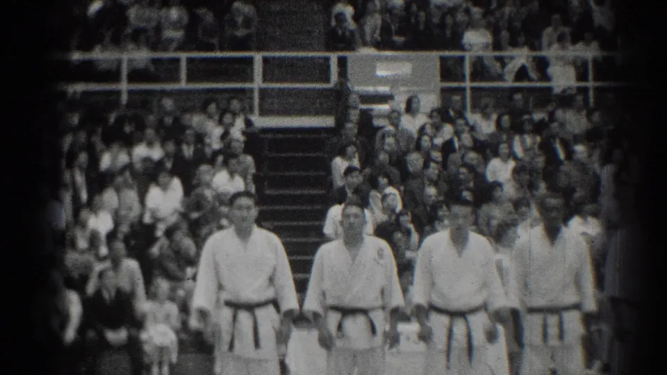 Four martial arts competitors in white gi and dark belts walk forward at the 1966 San Gabriel event, with bleachers behind them.