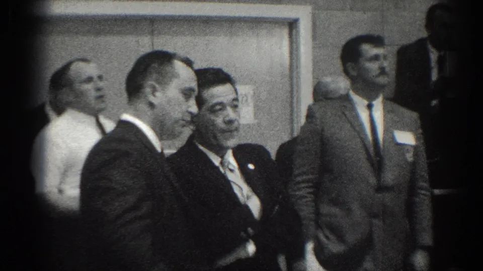 Suited officials or attendees stand near a doorway at the San Gabriel martial arts competition, filmed in black-and-white 8mm.