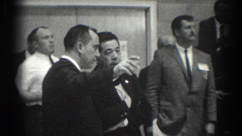 In San Gabriel, California, 1966, suited men converse in a hallway while other attendees stand nearby.