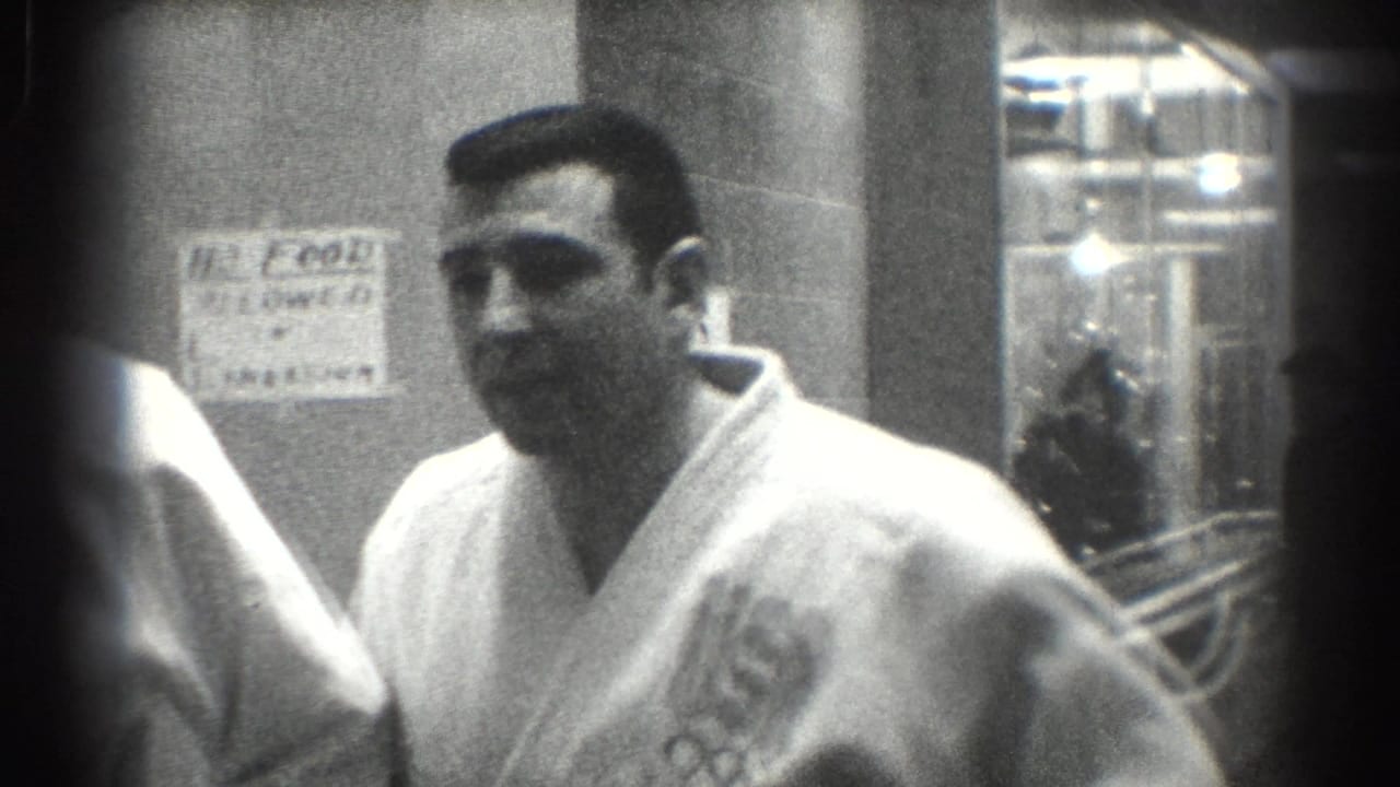 A man in a white gi stands in a hallway with a USA Olympic-ring patch visible.