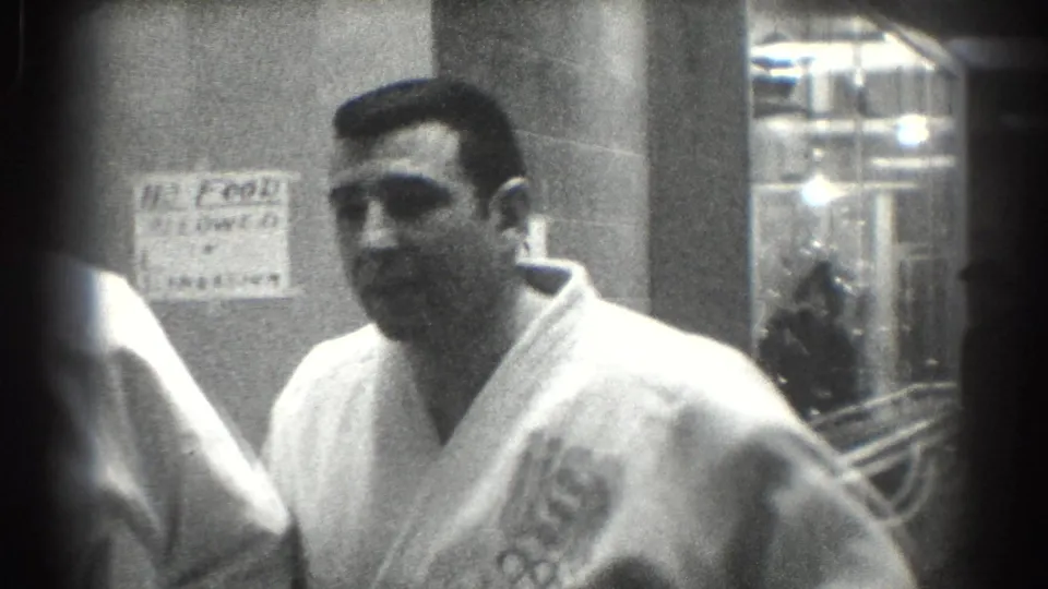 A karate team member in a white gi with a USA Olympic-ring patch stands in a San Gabriel hallway in 1966.