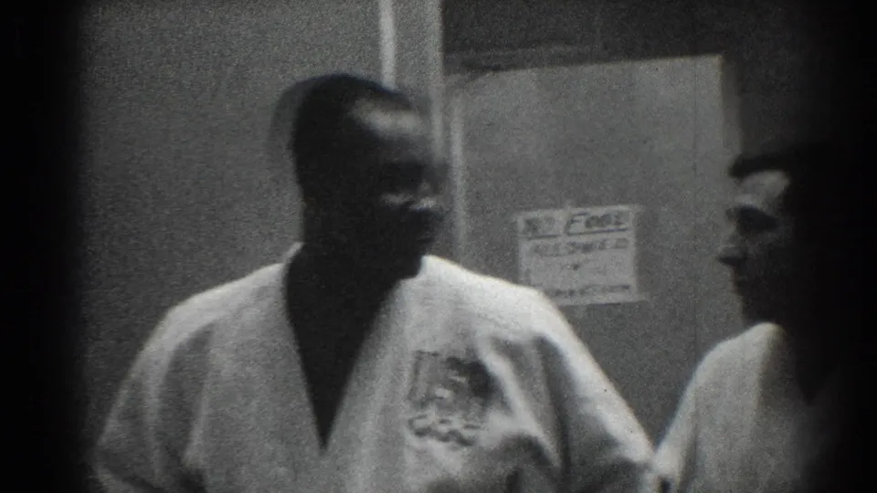 Two karate team members in white gis appear in a 1966 San Gabriel hallway, with one figure partly obscured by dark film edging.