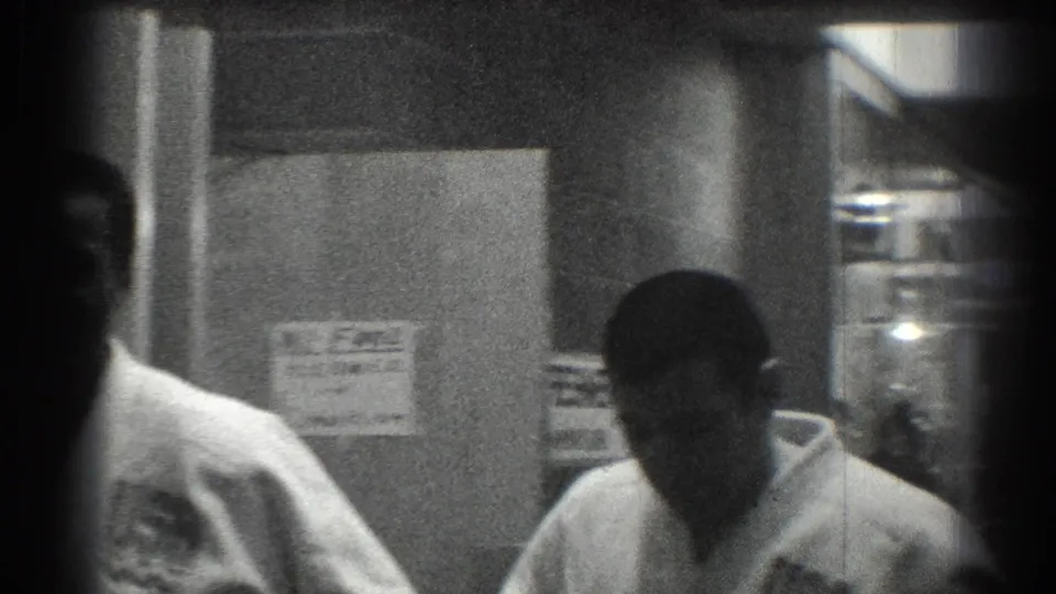 A karate team member in a white gi is seen near posted notices in a San Gabriel hallway, captured with soft 8mm framing.