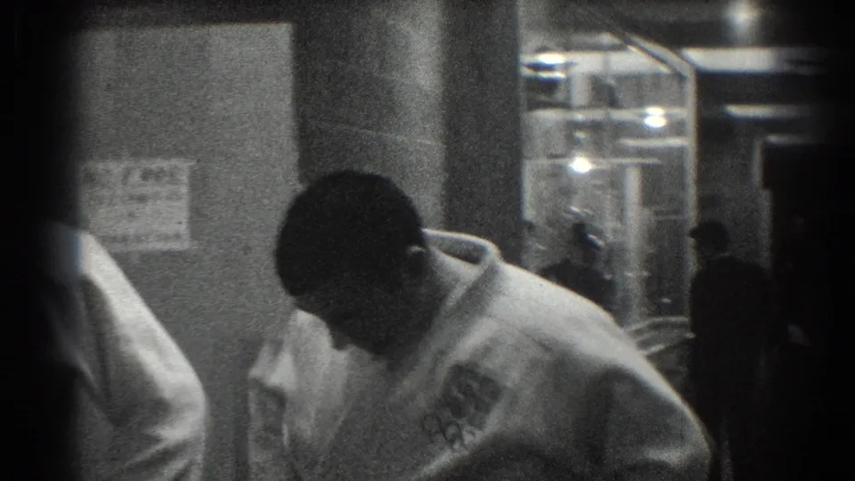 In San Gabriel, California 1966 footage, a martial artist in a white uniform bends forward near posted notices in a backstage area.