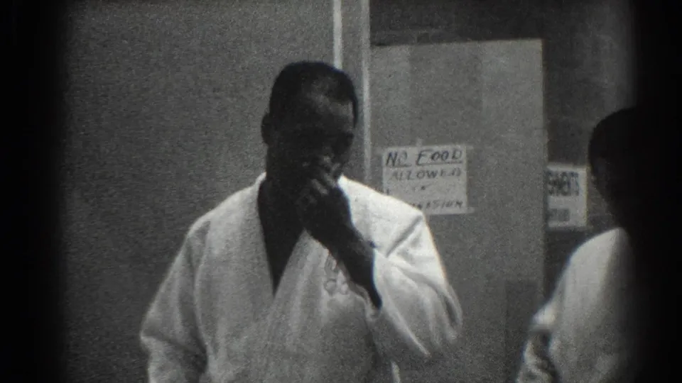 A man in a white martial arts uniform appears to smoke backstage during the 1966 San Gabriel event, with a no-food notice behind him.