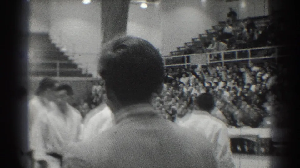 The camera looks from behind uniformed participants toward a crowded gymnasium during the 1966 San Gabriel martial arts event.