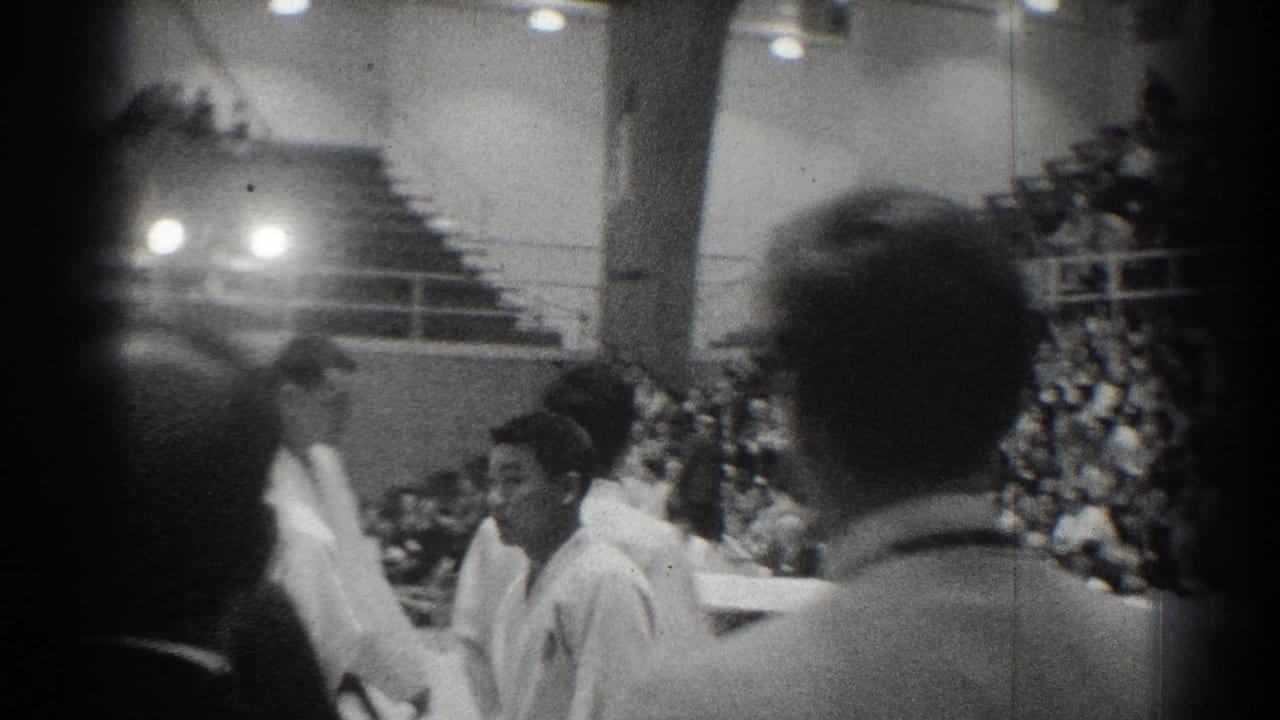 Men in white judo uniforms move through an indoor gym as spectators watch from bleachers.