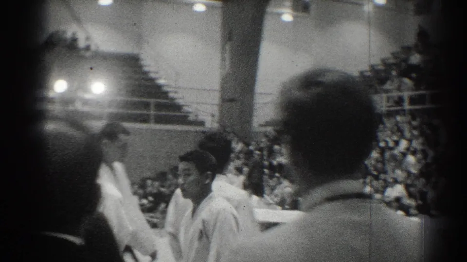 1966 San Gabriel gym scene with white-uniformed judo competitors passing in front of crowded bleachers and foreground spectators.