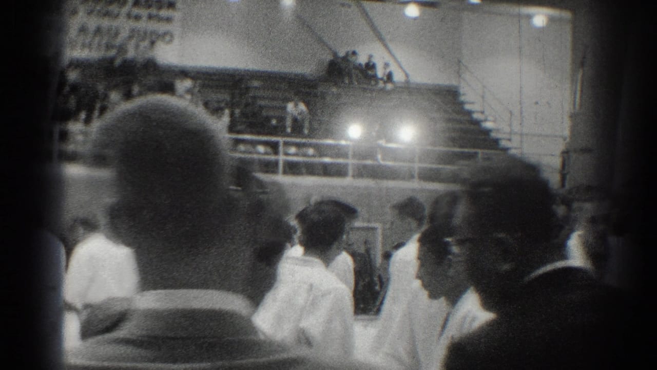Judo competitors and spectators are seen inside a gym, partly blocked by foreground onlookers.