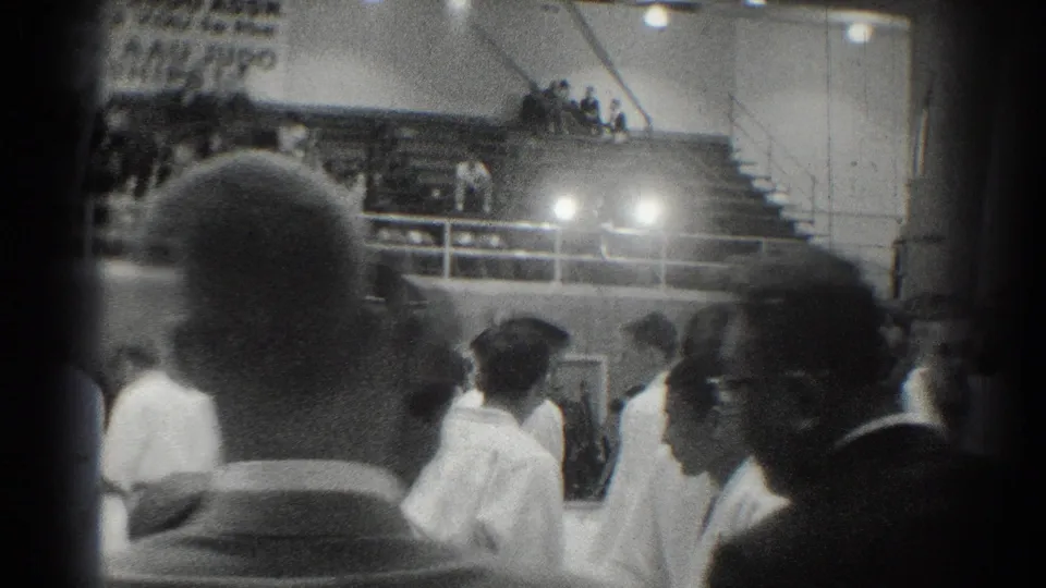 White-uniformed competitors gather in a San Gabriel gymnasium, with spectators in bleachers and foreground heads obscuring part of the view.