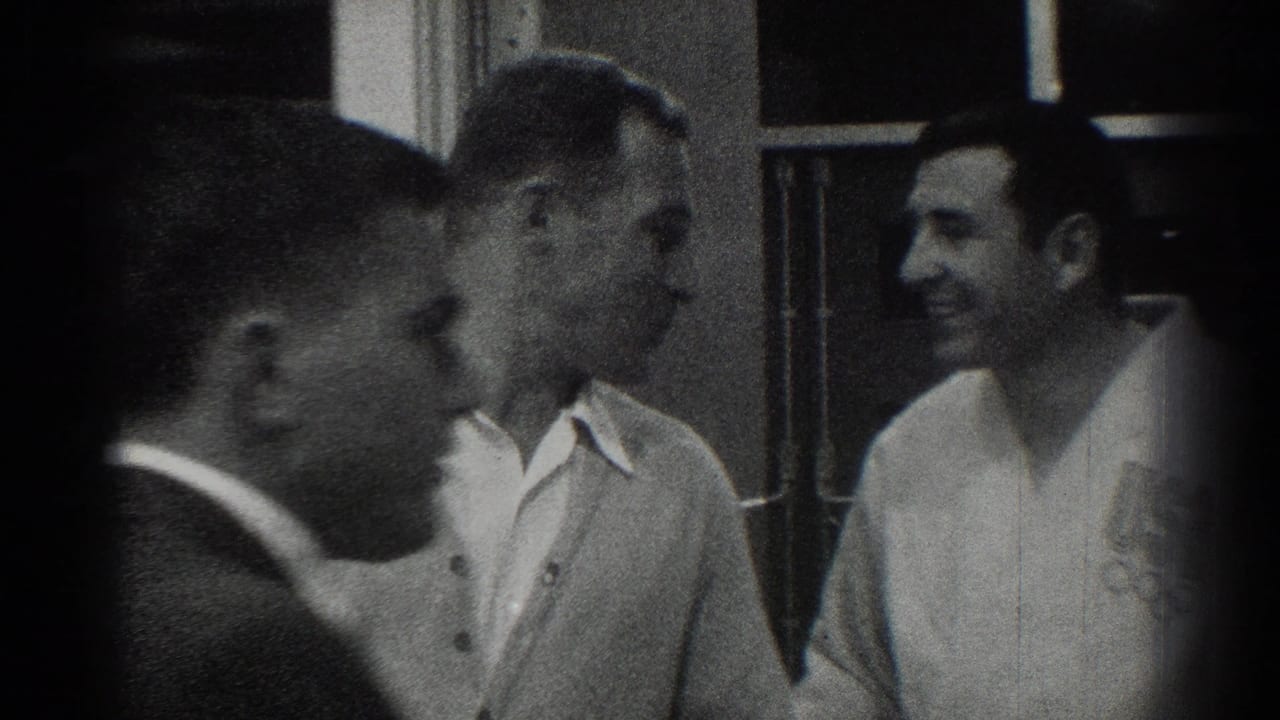 Men in white uniforms gather indoors while a man in a dark jacket stands close to the camera.