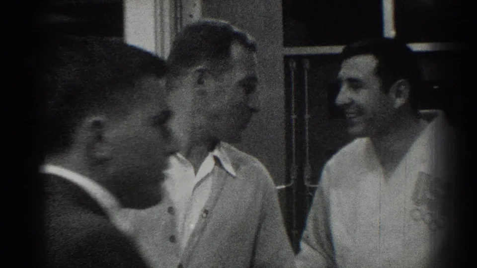 1966 San Gabriel home-movie frame showing adult men in white martial arts-style uniforms near interior windows, with a dark-jacketed man in the foreground.