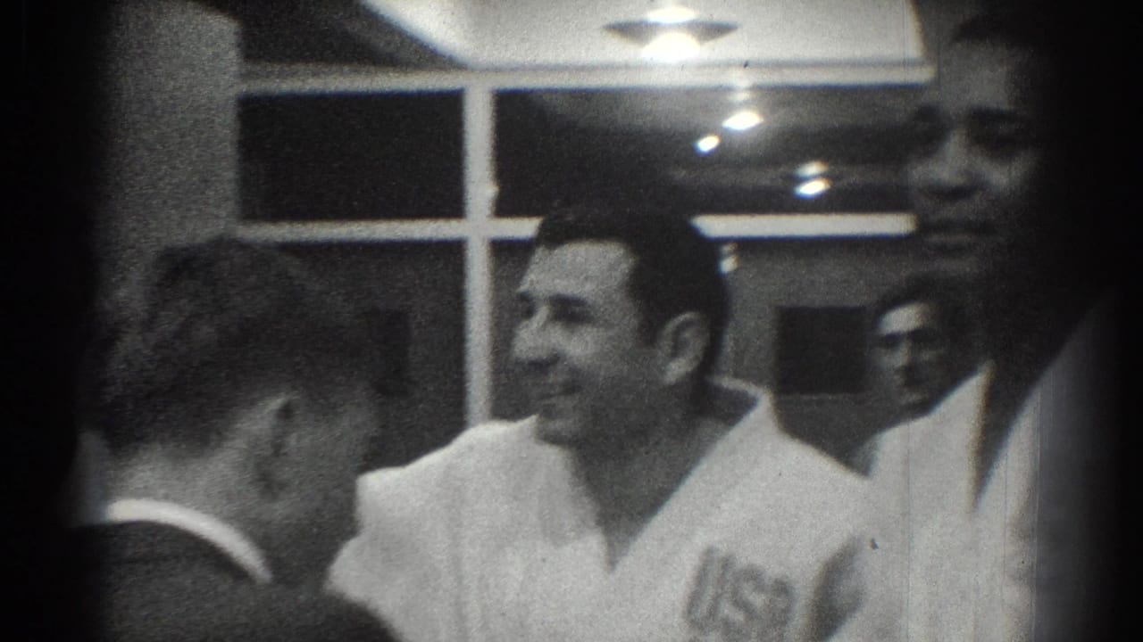 1966 San Gabriel frame of a smiling adult man in a white martial arts-style uniform among other men inside a training-room-like interior.
