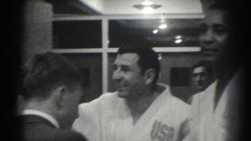 1966 San Gabriel frame of a smiling adult man in a white martial arts-style uniform among other men inside a training-room-like interior.