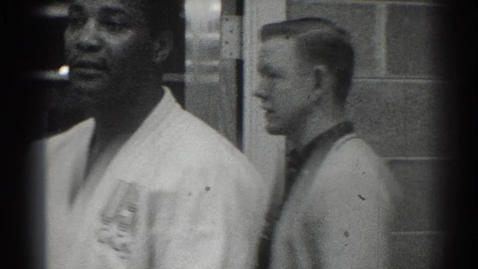 1966 San Gabriel home-movie frame with a white-uniformed adult man in close foreground and another man standing by the interior wall.