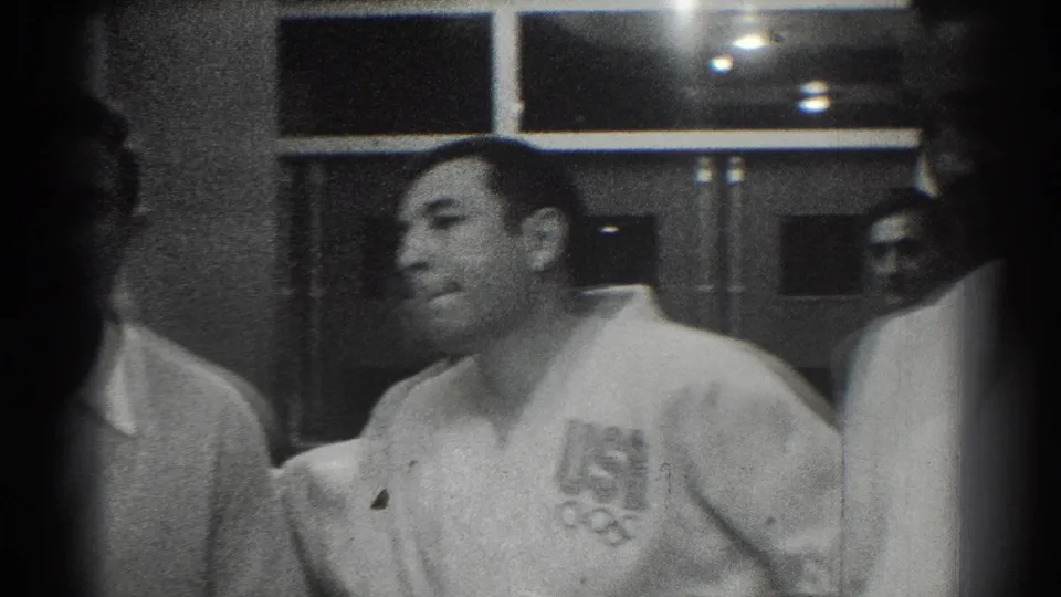 1966 San Gabriel frame showing a smiling adult man in a white martial arts-style uniform beside others in a softly lit interior.