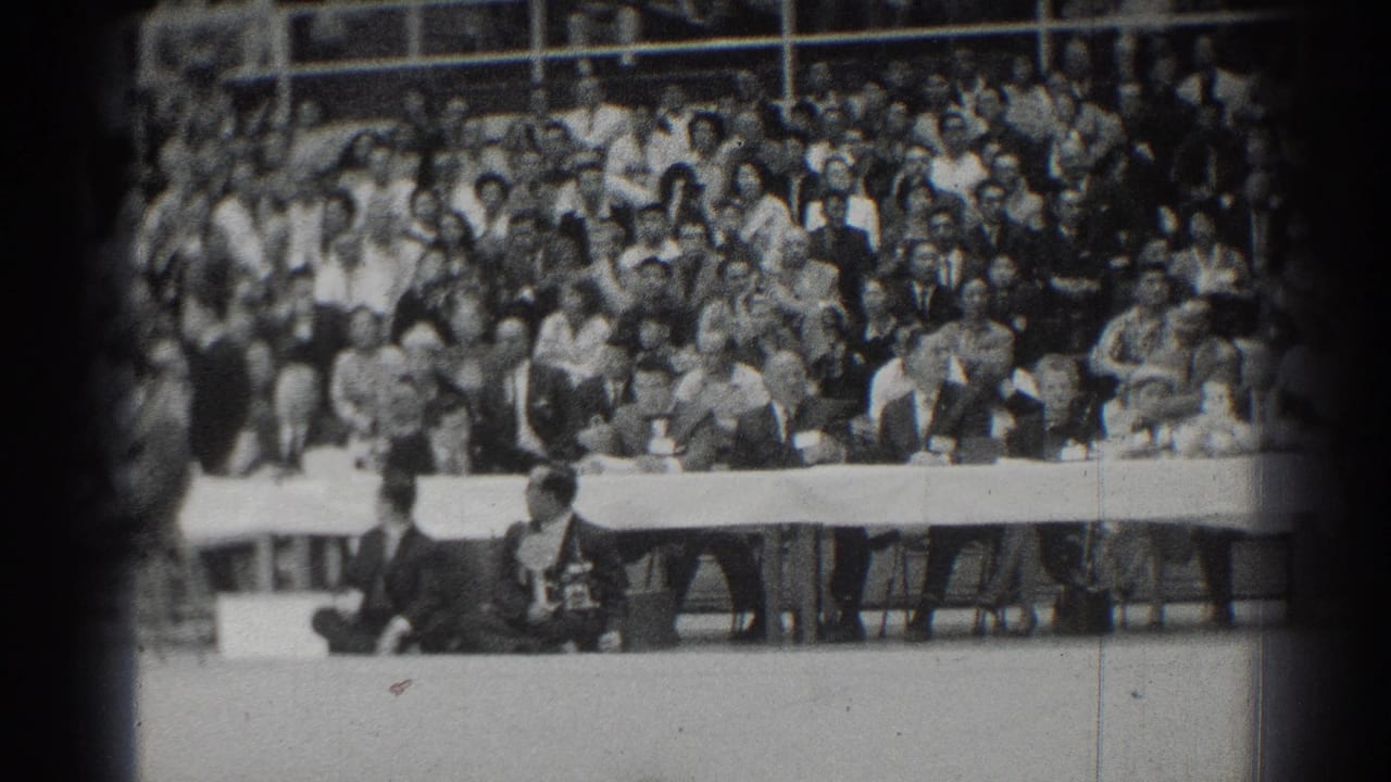 Crowd seated in bleachers behind officials at a long table during an indoor martial arts event.