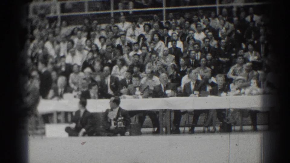 San Gabriel 1966 home-movie frame showing spectators in bleachers and suited officials seated at a long table during a karate match.