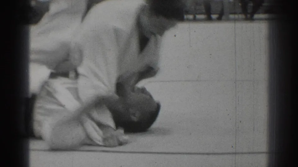 Black-and-white 8mm frame from a 1966 San Gabriel karate match showing martial artists on the gym floor in close action.