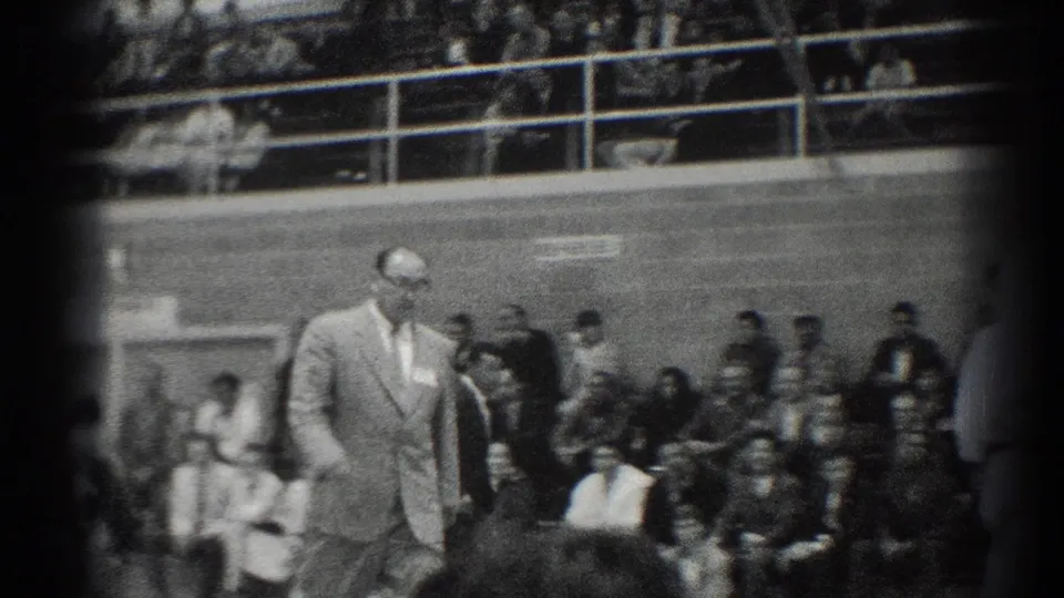 San Gabriel 1966 frame showing a suited man, likely an event official, walking past spectators during an indoor karate match.