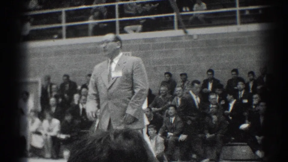 Black-and-white 8mm frame from the San Gabriel karate event showing a suited official in front of a seated audience.