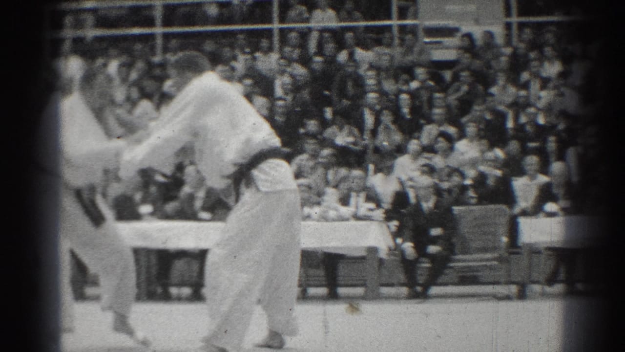 Two judo competitors in white gi grapple on a mat before seated spectators.