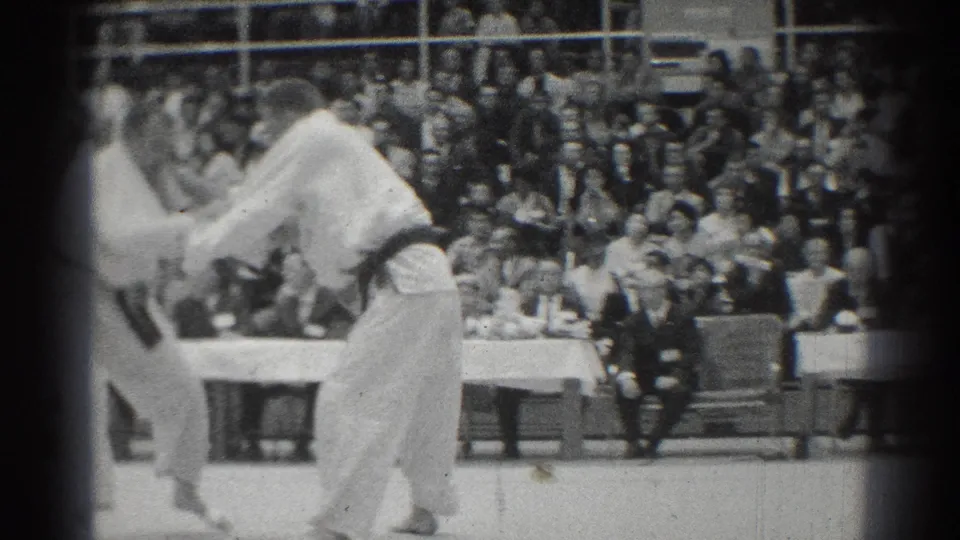 Two judo competitors in white gi and black belts grapple during a 1966 San Gabriel match as a crowd watches from bleachers.