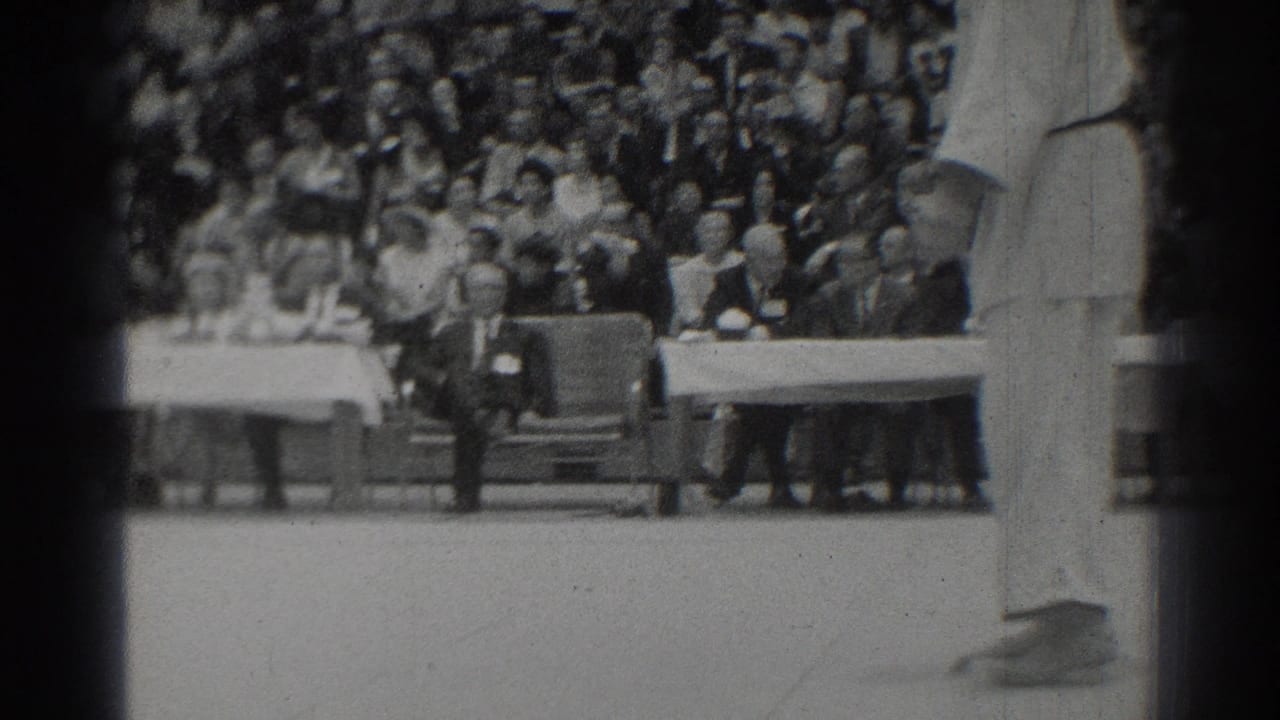 A suited official stands near tables with a large audience seated behind him.