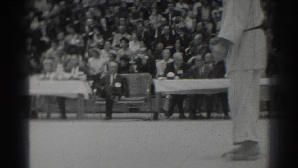 A suited official stands beside long tables during the San Gabriel judo event, with spectators packed into bleachers behind him.