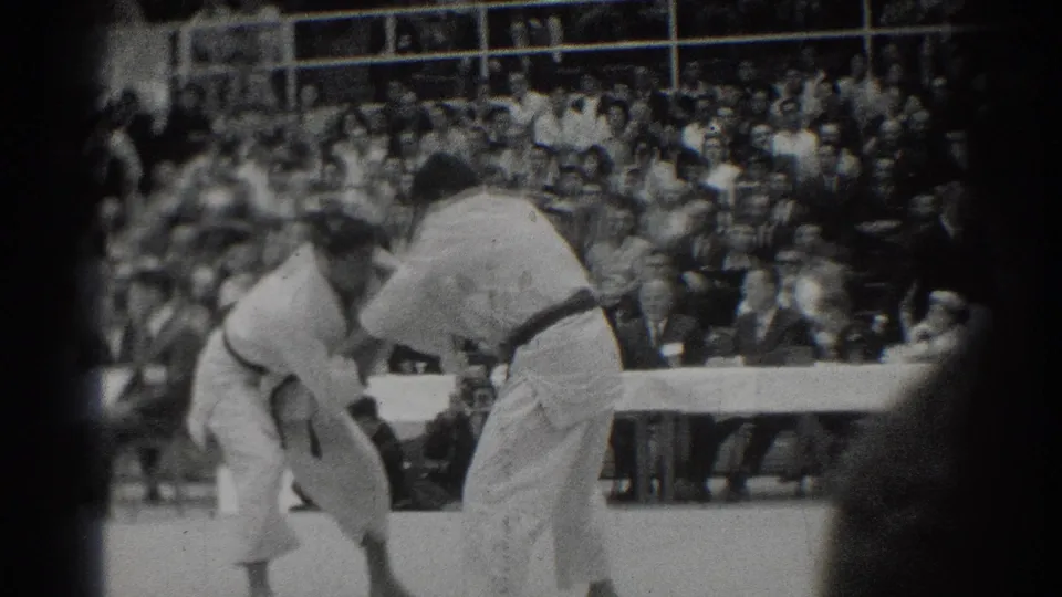 Two competitors in white gi lean into a grip exchange during the 1966 San Gabriel judo match, filmed from the spectator side.