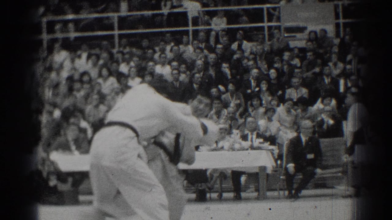 Two judo competitors in white gi grapple on a mat before a crowded gym audience.