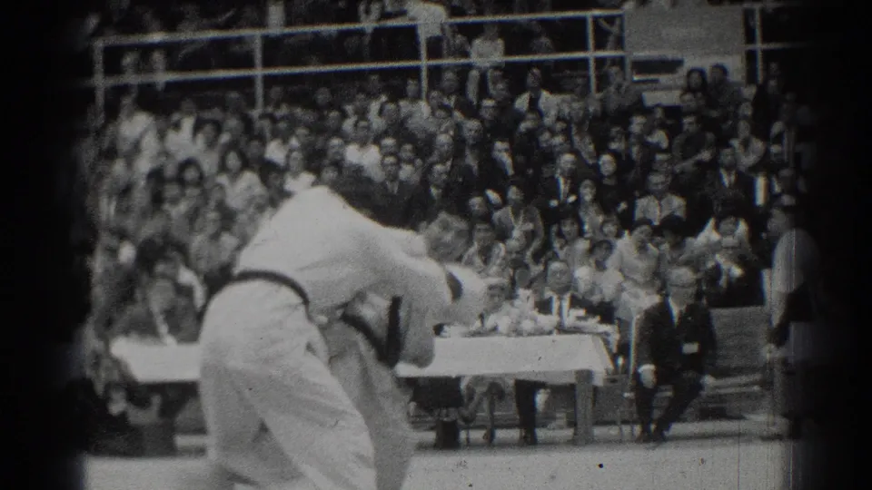 In 1966 San Gabriel footage, two judo competitors in white gi grapple on a mat while spectators and officials watch from the gymnasium.