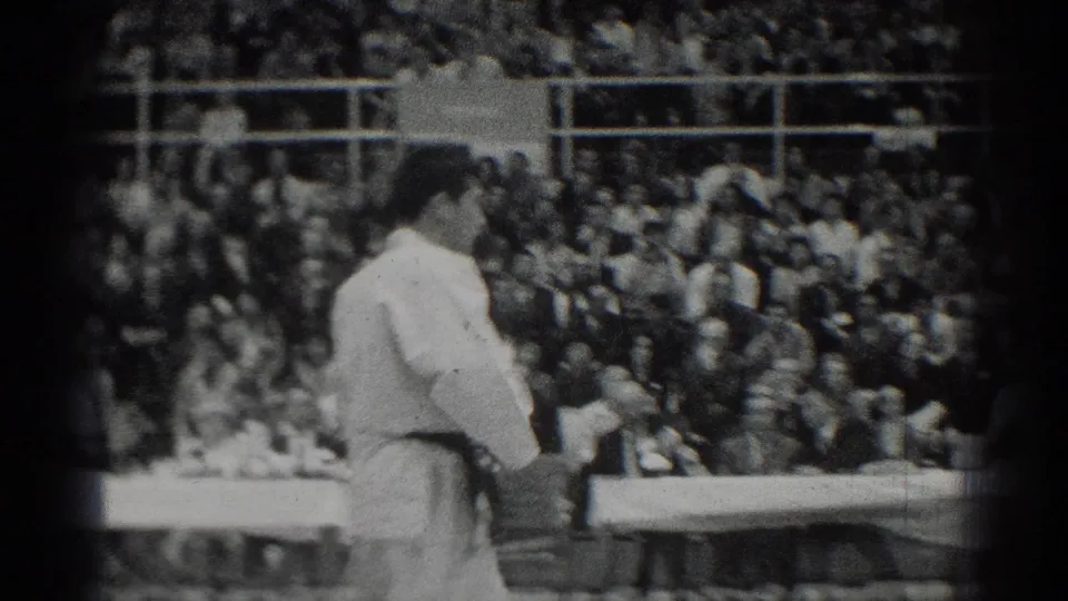 One judo competitor in a white gi stands in profile near the mat during the San Gabriel match, with judges and audience members visible behind him.