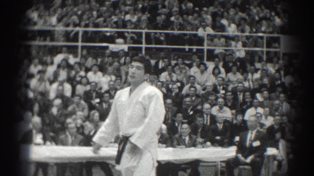 A karate competitor in a white gi stands during a San Gabriel, California match, with spectators filling the bleachers behind him.