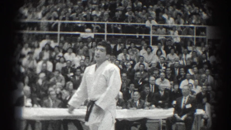 A karate competitor in a white gi stands during a San Gabriel, California match, with spectators filling the bleachers behind him.