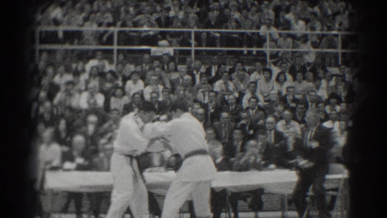 Two karate competitors grapple on a platform while a referee stands nearby.