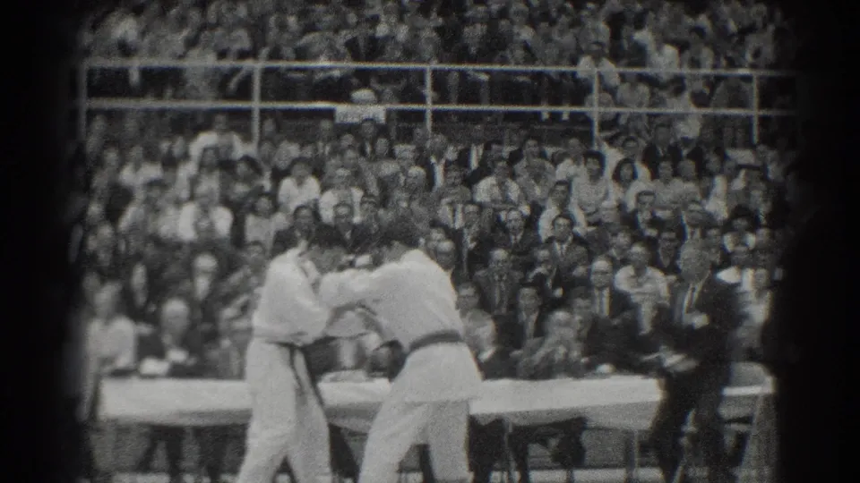 Two competitors in white gi engage during the San Gabriel karate match as a suited referee watches beside the platform.